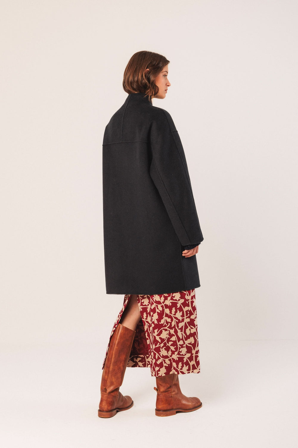 Manteau minimaliste à coupe ample, col montant, porté sur une robe longue avec des bottes en cuir.
