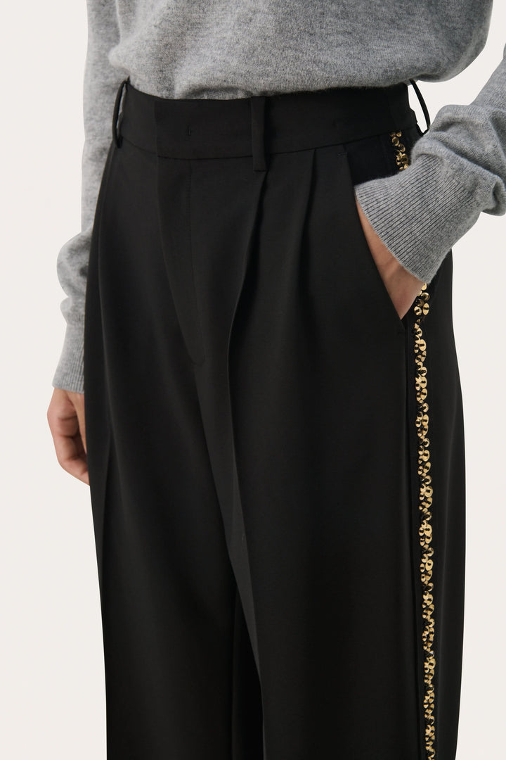 Pantalon noir large avec bande brodée dorée, taille haute et plis subtils, offrant un look élégant et raffiné.