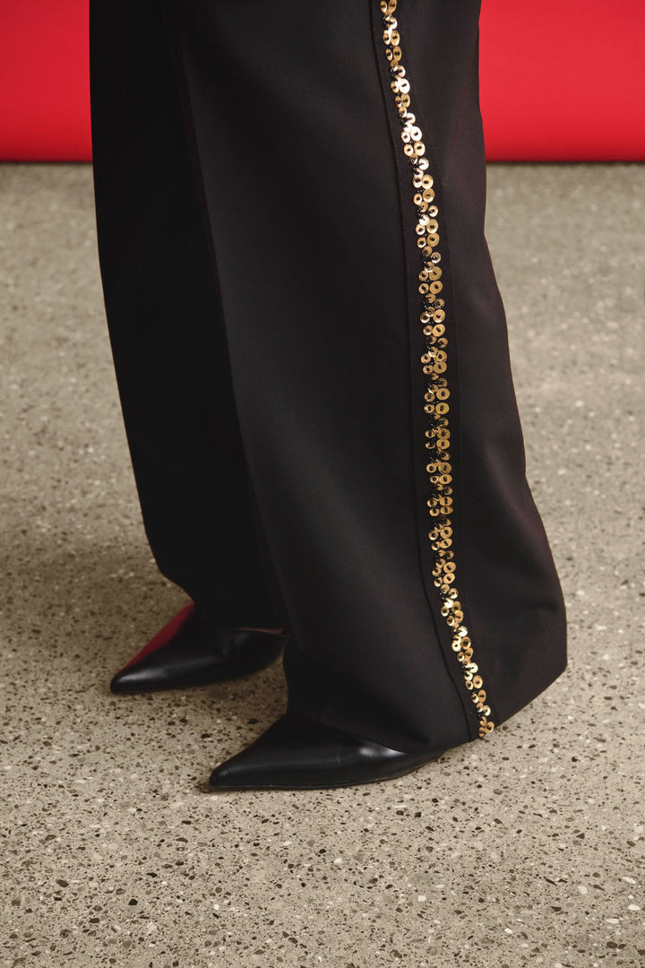 Pantalon noir large avec bande brodée dorée sur le côté, porté avec des chaussures noires élégantes.