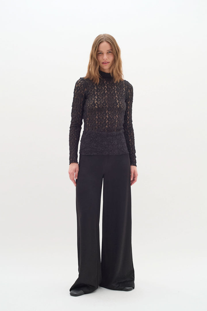 Femme portant un pantalon satiné noir et un haut en dentelle, style chic et moderne, sur fond blanc.