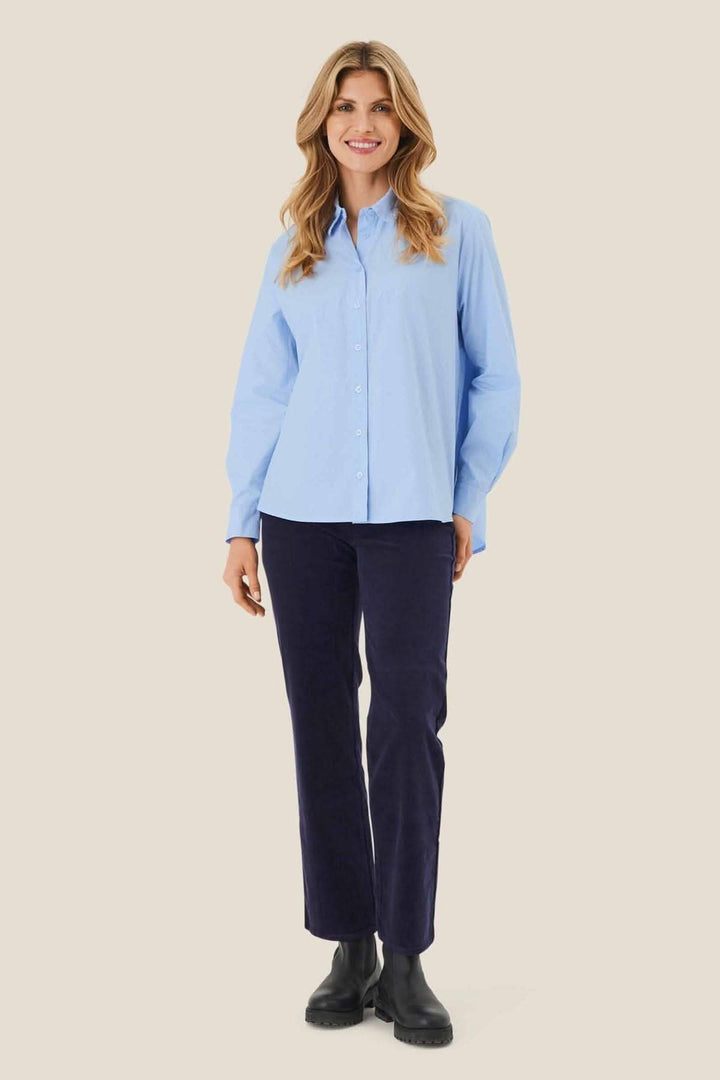 Femme souriante portant une chemise bleue et un pantalon noir, affichant un look élégant et décontracté.