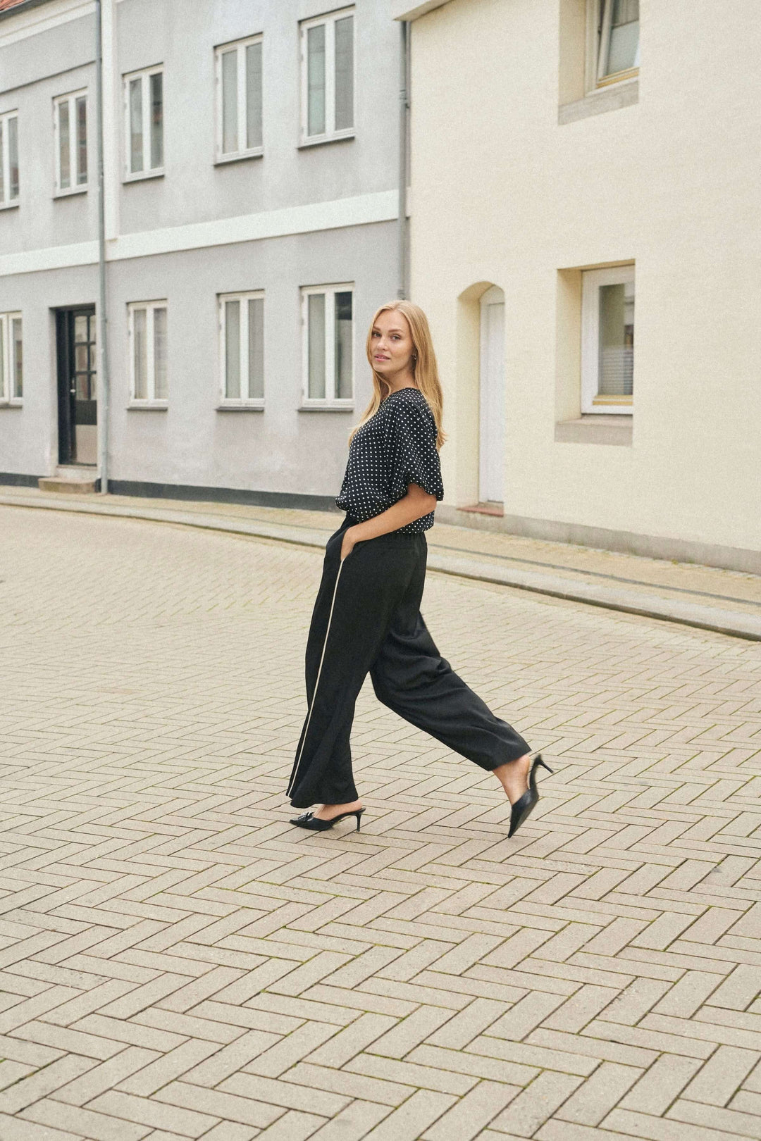 Femme marchant dans un pantalon fluide noir à jambe large et un haut à pois, dans une rue pavée.