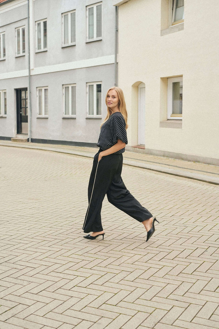 Femme marchant dans un pantalon fluide noir à jambe large et un haut à pois, dans une rue pavée.