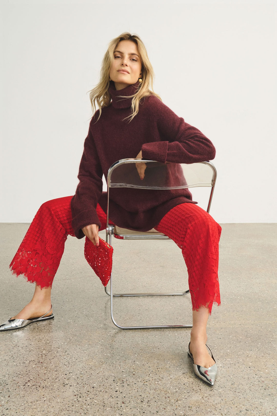 Femme élégante portant un pantalon en dentelle rouge et un pull à col roulé, assise sur une chaise.