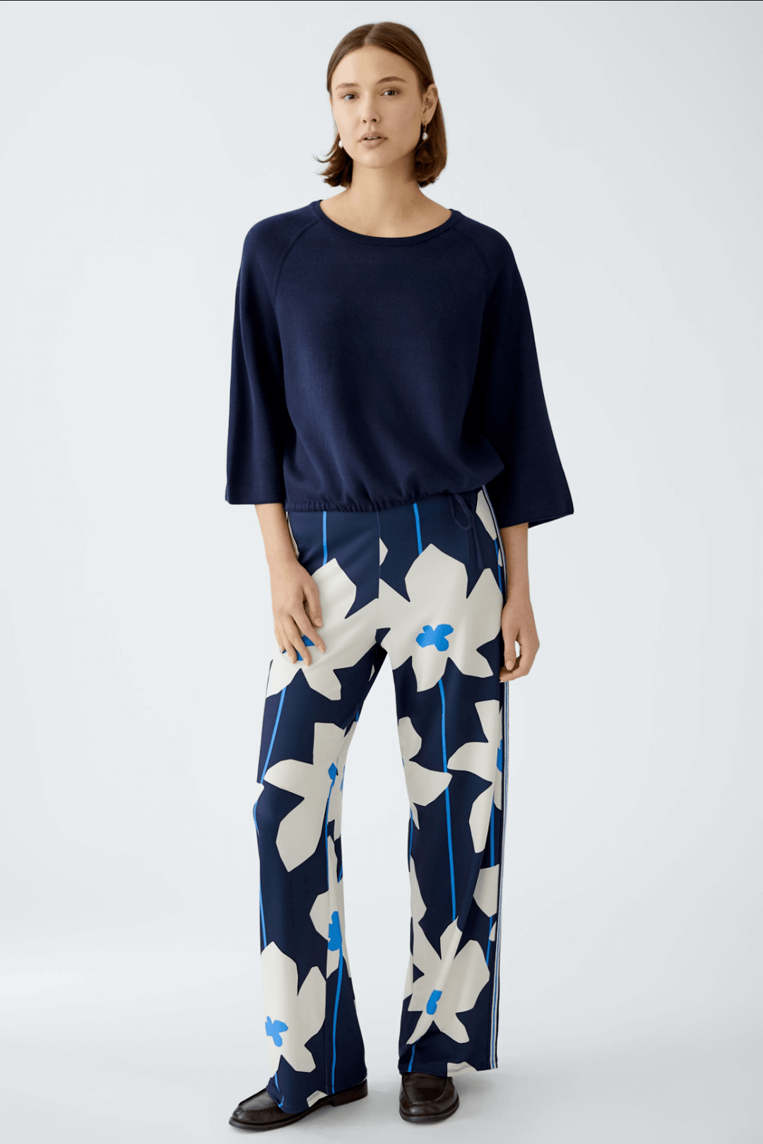 Femme portant un pantalon imprimé fleurs bleu et blanc avec un pull noir, style chic et décontracté.