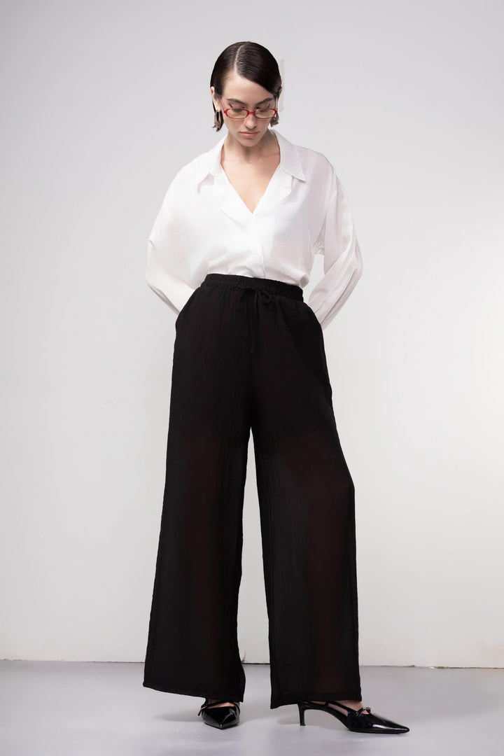 Femme portant un pantalon jambe large noir avec blouse blanche, offrant une silhouette fluide et moderne, taille élastique.