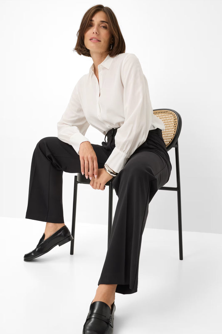 Femme assise sur une chaise, portant un pantalon palazzo noir et une chemise blanche, style chic et élégant.
