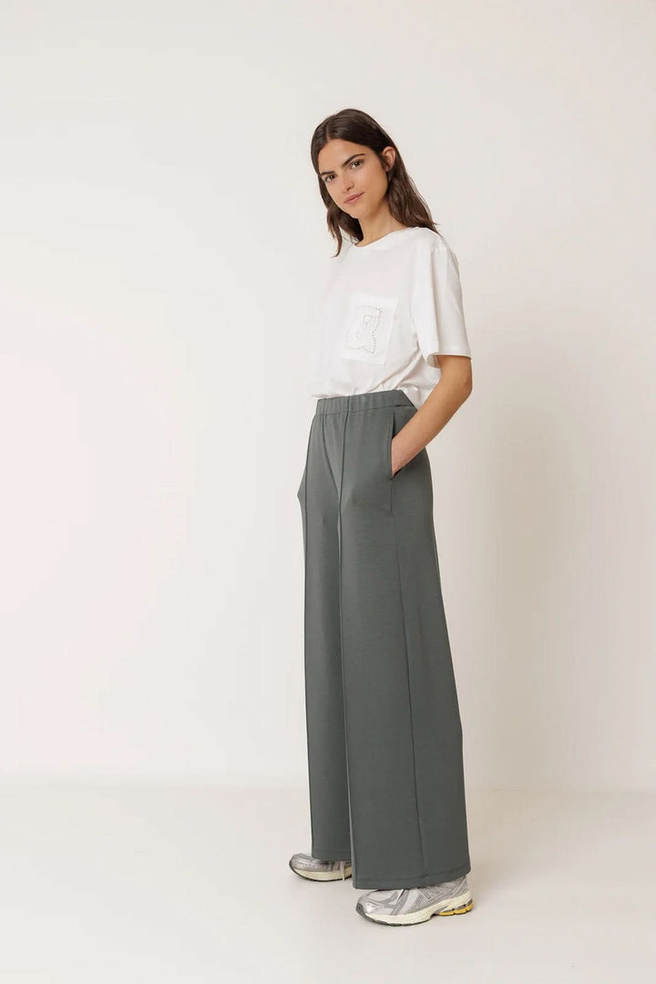 Femme portant un pantalon tailleur fluide à plis marqués avec un t-shirt blanc et des baskets grises.