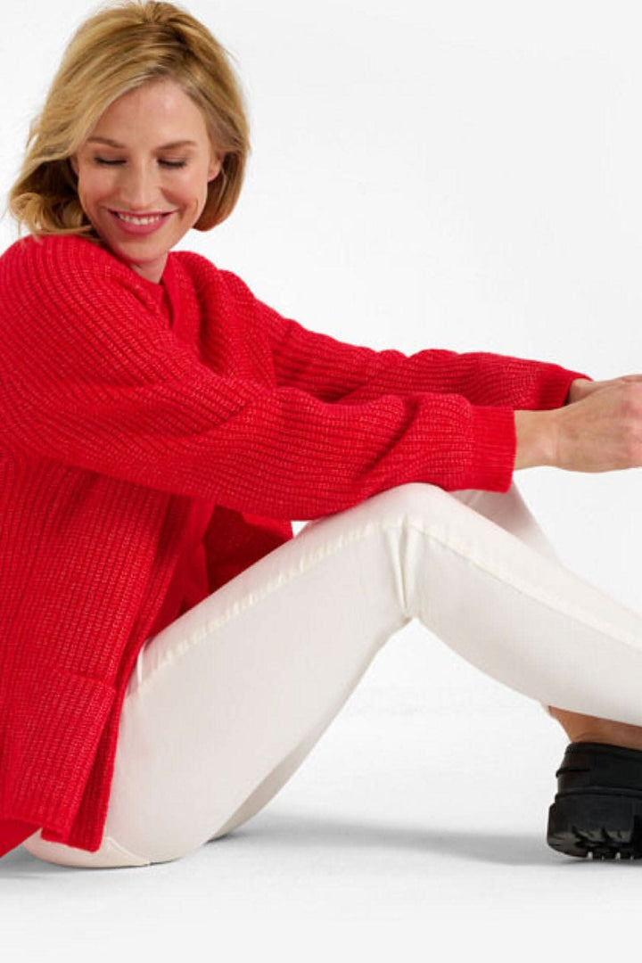 Femme souriante assise, portant un pull rouge et pantalons blancs, pour un look décontracté et élégant.