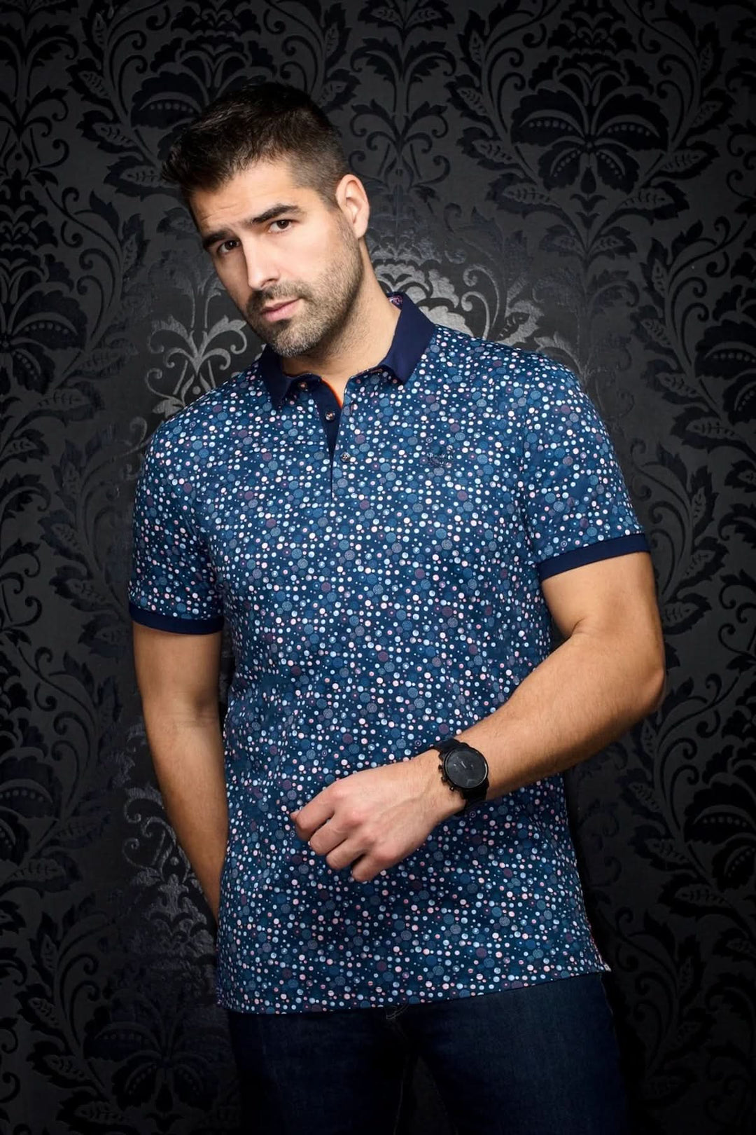 Homme portant un polo à imprimé à pois multicolores sur fond marine, col contrastant, style décontracté chic.