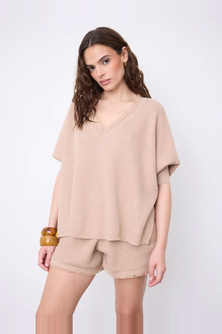 Model wearing Pull poncho col V à manches ¾ in beige cotton cashmere with fringed edges and side slits for a modern look.
