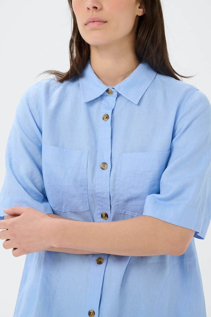 Femme portant une robe chemise en lin Sardiae bleue avec manches courtes et fermeture boutonnée, idéal pour un look décontracté et soigné.