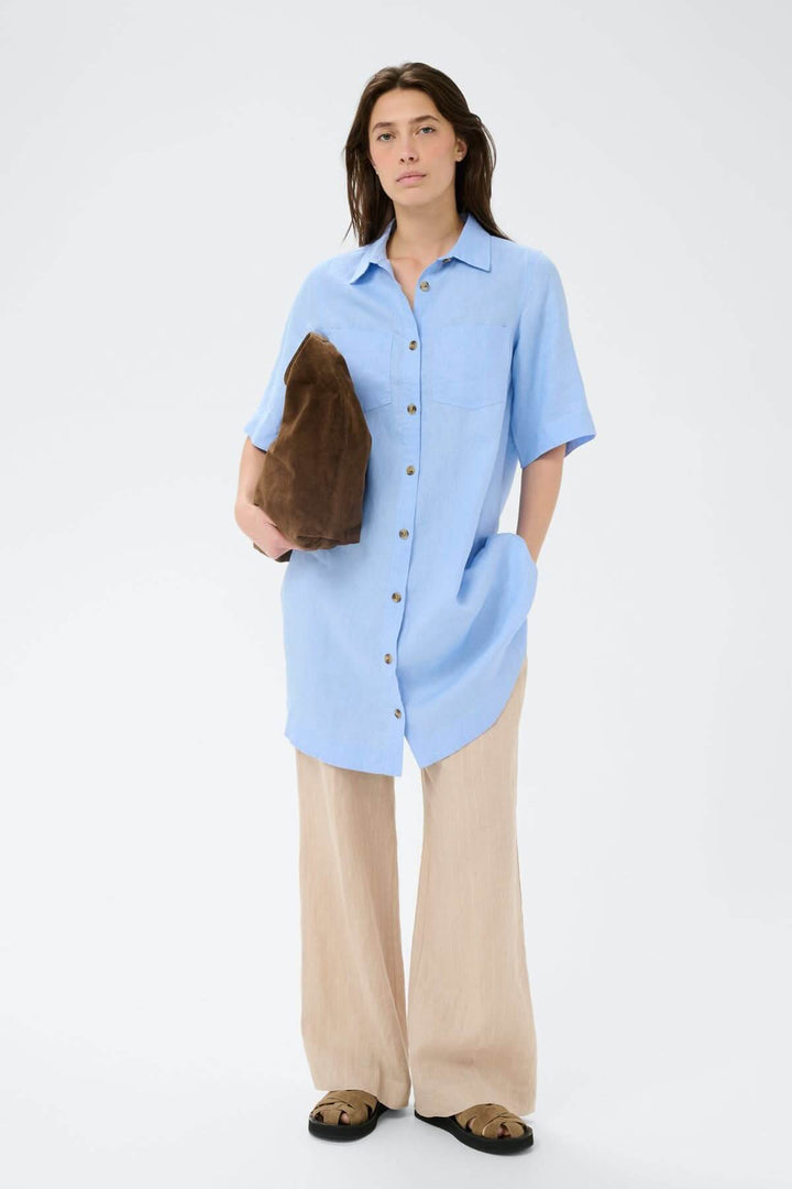 Femme portant une robe chemise en lin Sardiae bleue avec fermeture boutonnée, style décontracté, urbain et minimaliste.