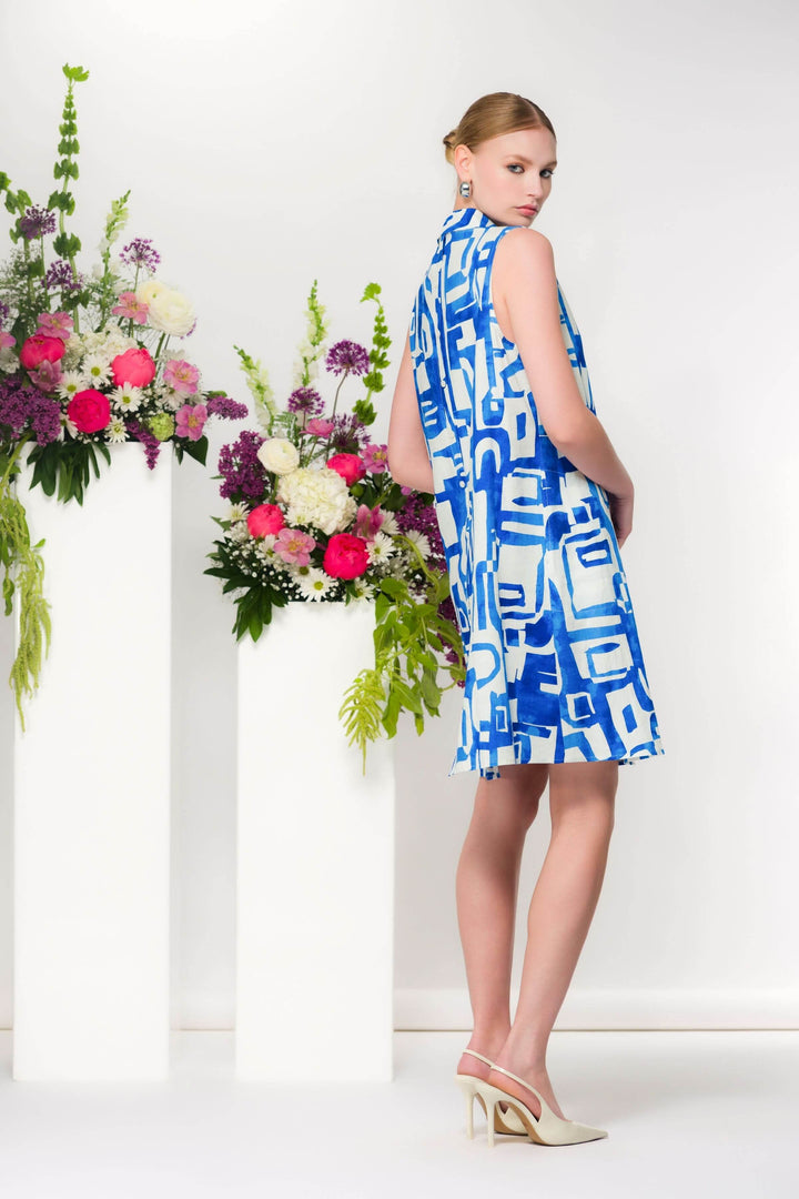 Robe col chemisier avec imprimé graphique bleu et blanc, silhouette fluide et évasée, sans manches, devant un bouquet de fleurs colorées.