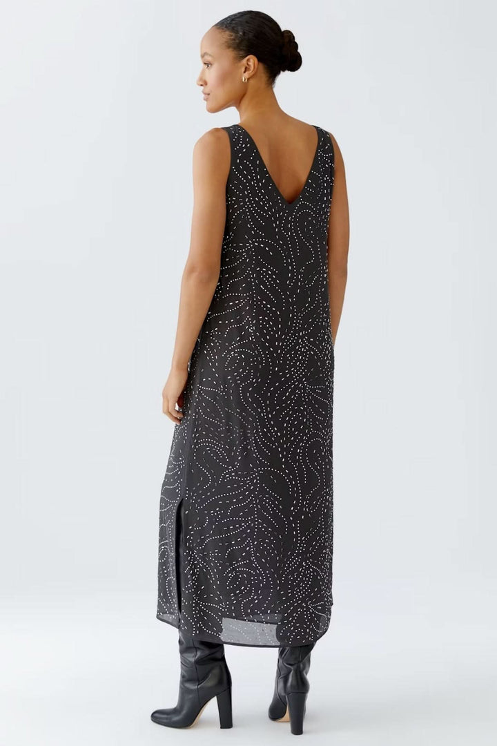 Robe longue sans manches avec imprimé discret en pointillés, encolure en V, fentes latérales, portée avec des bottes hautes.