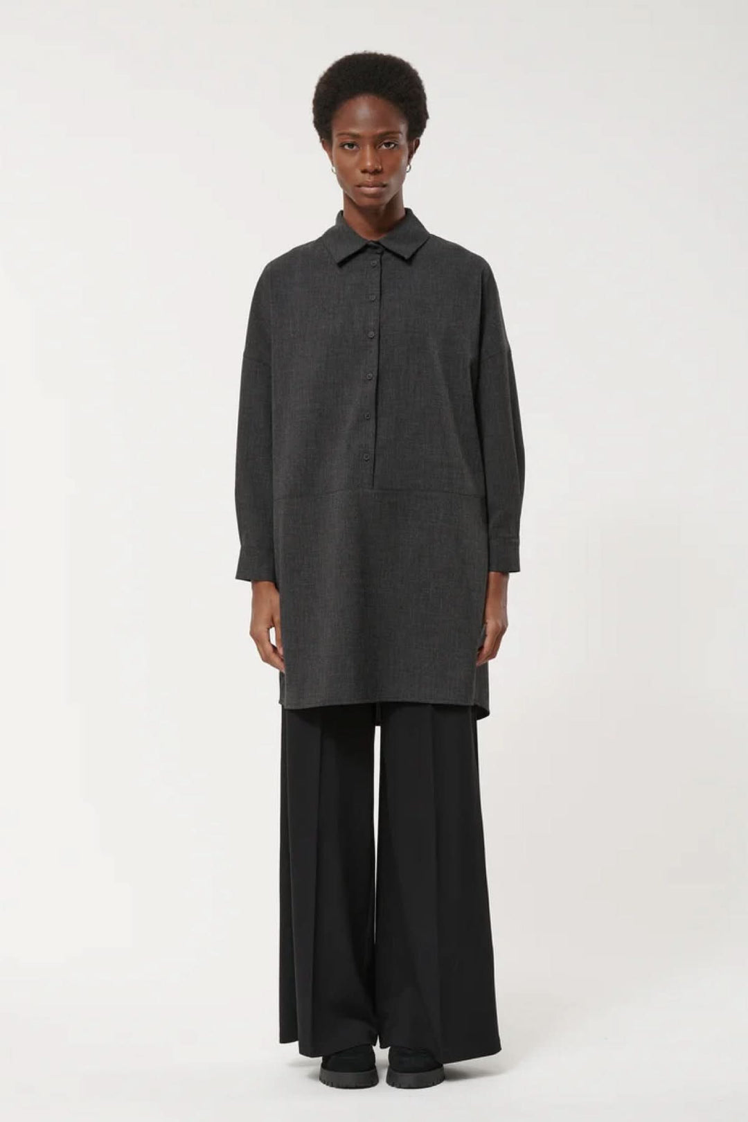 Femme portant une robe à manches longues, coupe ample et design minimaliste, avec col classique et fermeture boutonnée. Style moderne et épuré.