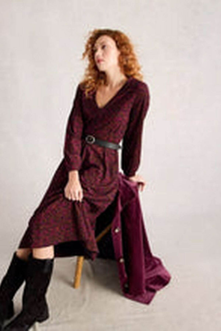 Femme portant une robe midi en jersey col en V avec manches longues, jupe évasée, ceinture noire et bottes hautes, assise sur une chaise.