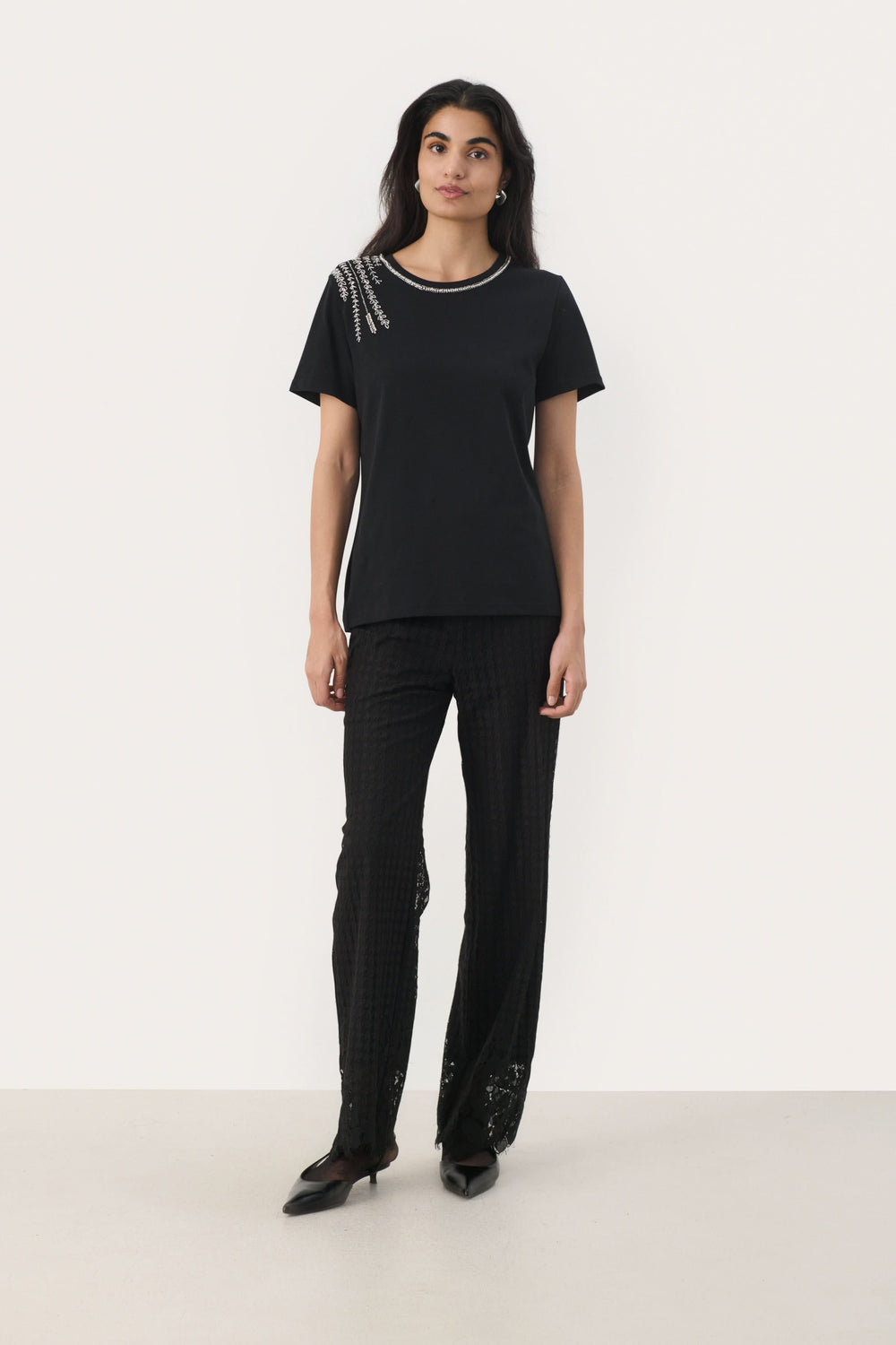 T-shirt noir à col rond avec broderies argentées et détails en perles, porté avec un pantalon noir élégant.