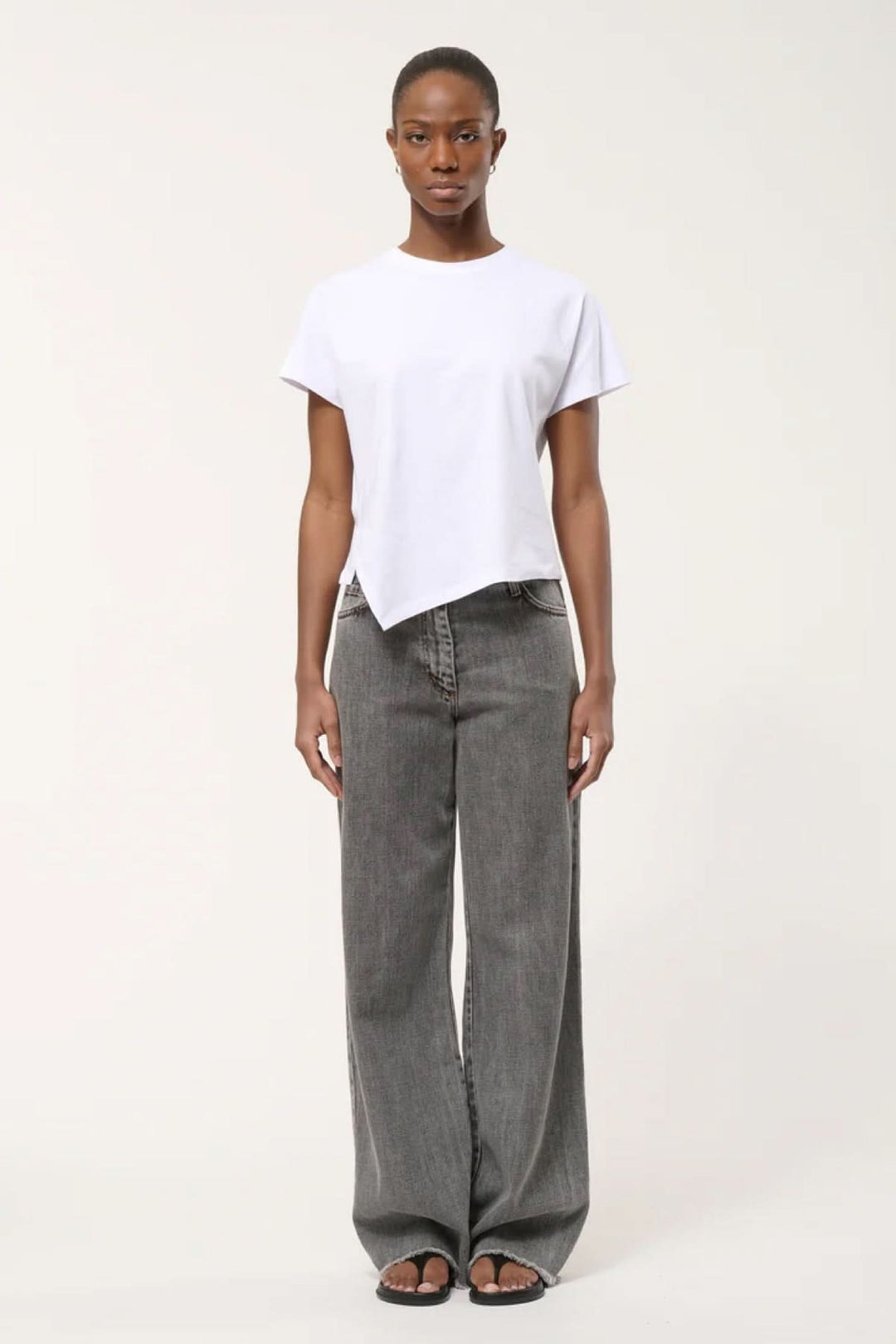 Femme portant un T-shirt à ourlet asymétrique blanc, style moderne et épuré, associé à un jean ample gris.