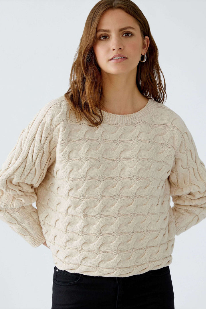 Femme portant le tricot Sirah, motif torsadé, coupe carrée moderne, coton doux et volumineux, couleur beige élégante.