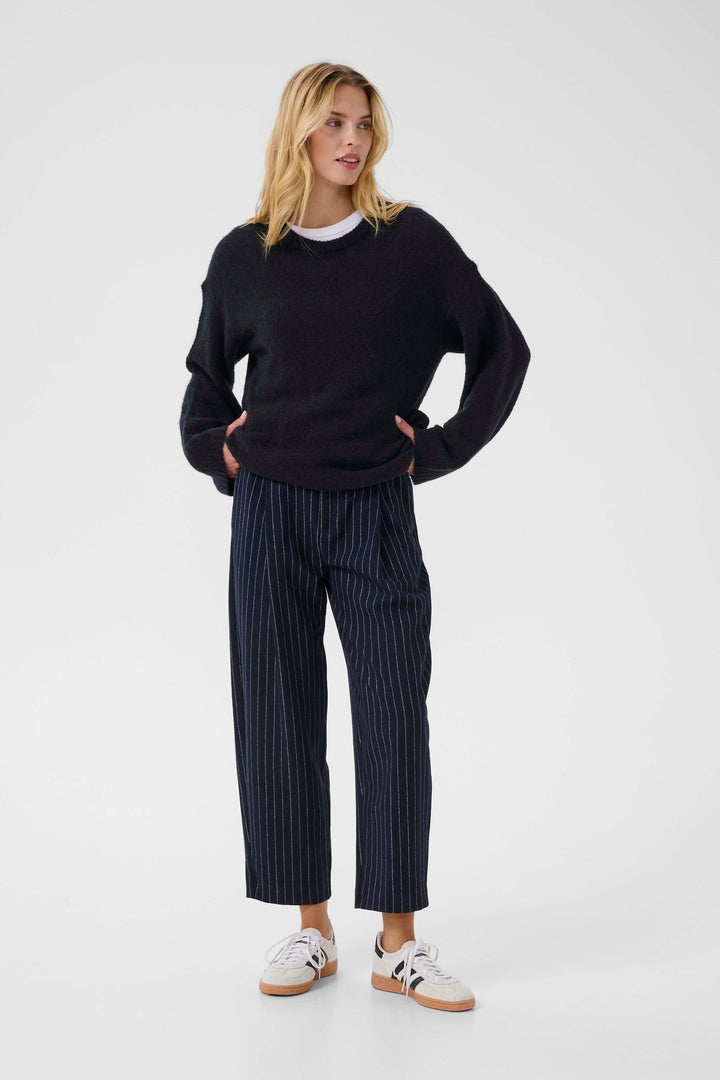 Femme portant un Tricot Spence noir à col rond, manches longues, avec pantalon à pinces rayé et baskets blanches.