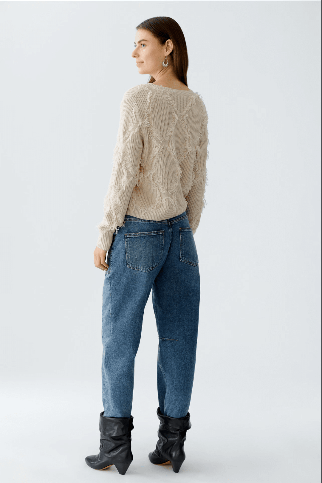 Femme portant tricot motif losange avec franges, coupe courte et détails métalliques, assorti à un jean taille haute.