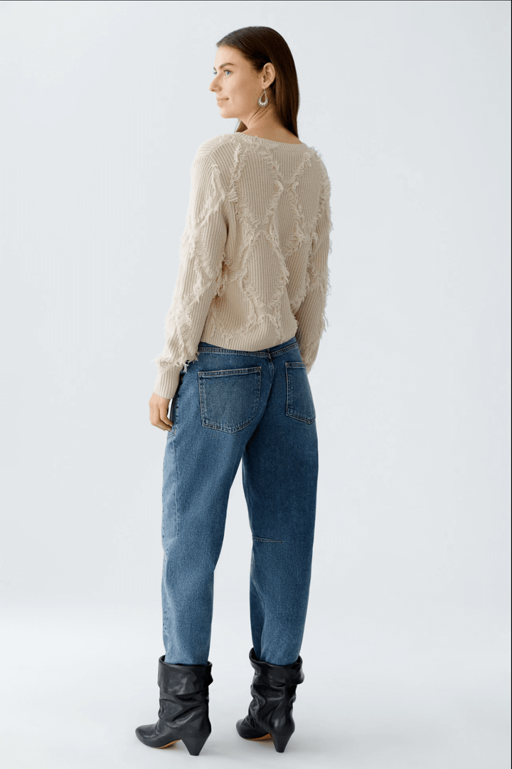 Femme portant tricot motif losange avec franges, coupe courte et détails métalliques, assorti à un jean taille haute.