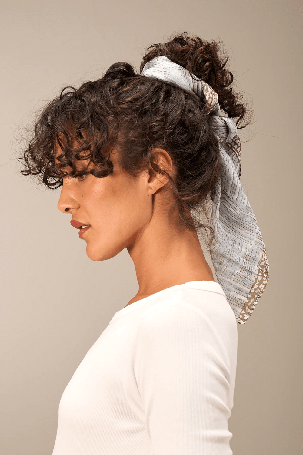 Femme portant un foulard imprimé élégant et raffiné noué dans ses cheveux bouclés, ajoutant une touche chic à sa tenue simple.