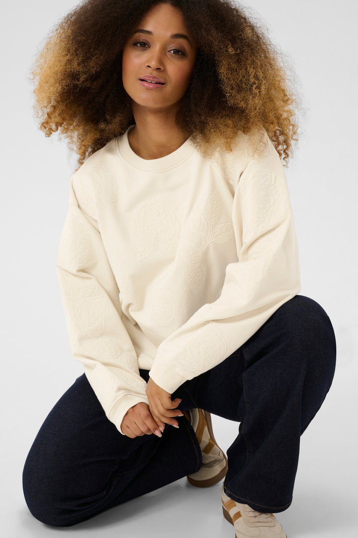 Femme portant un coton ouaté crème avec broderies florales, associé à un pantalon foncé pour un style élégant.