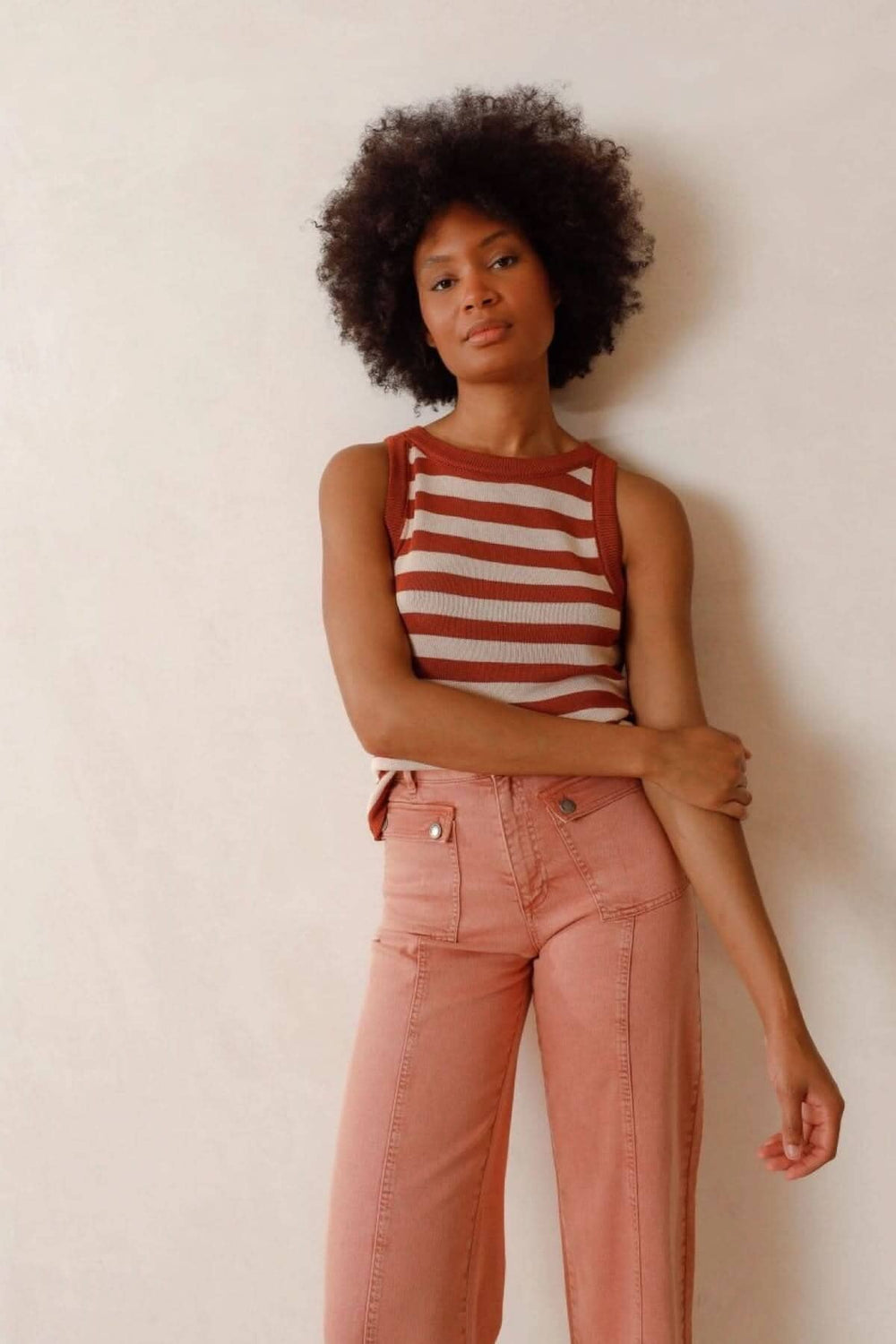 Femme avec un haut à rayures marron et crème, portant un pantalon rose, pose contre un mur neutre.