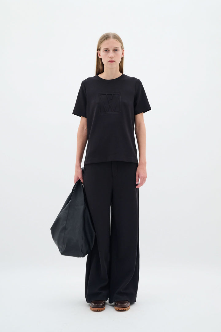 Femme portant un t-shirt noir et un pantalon large noir, tenant un sac noir, sur fond blanc.