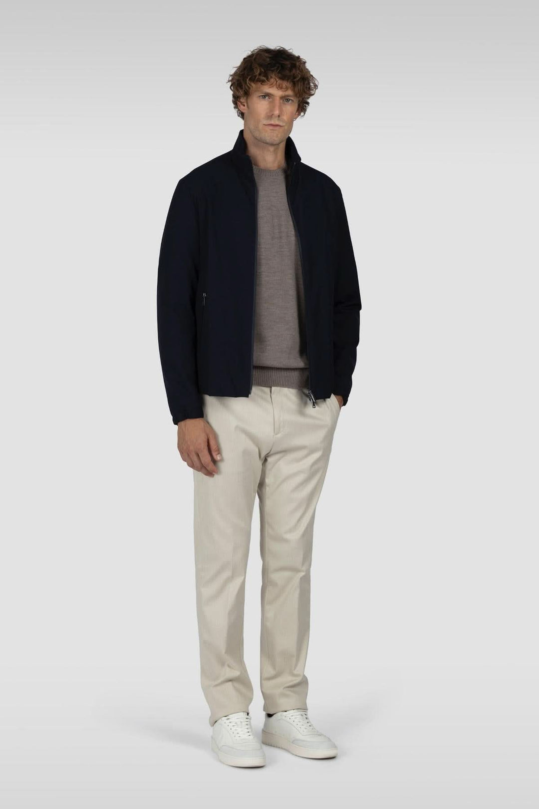 Homme debout en tenue élégante avec blouson noir, chandail gris et pantalon clair, chaussé de baskets blanches.