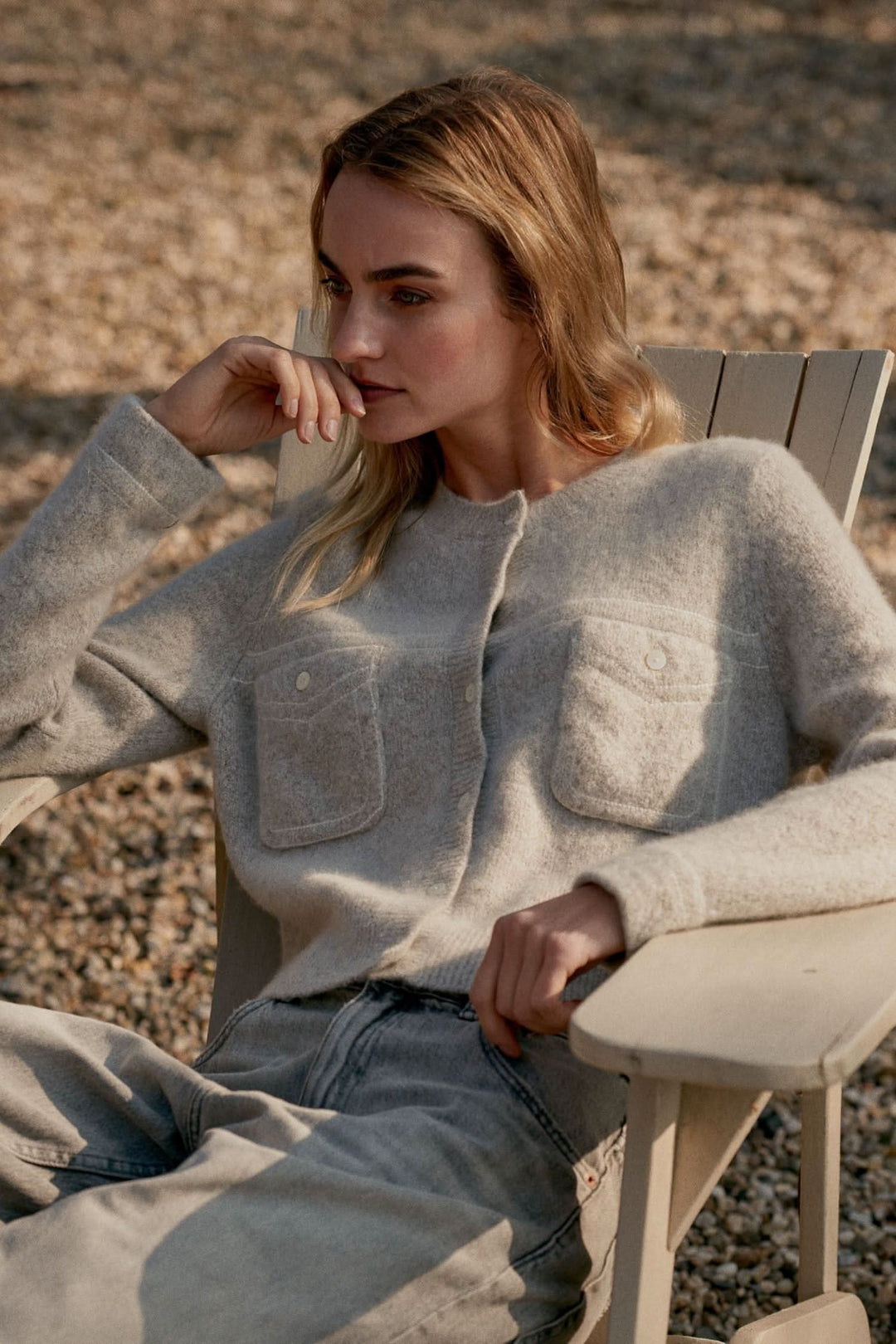 Femme portant un cardigan en maille doux avec poches plaquées, assise sur une chaise en bois, ambiance décontractée.