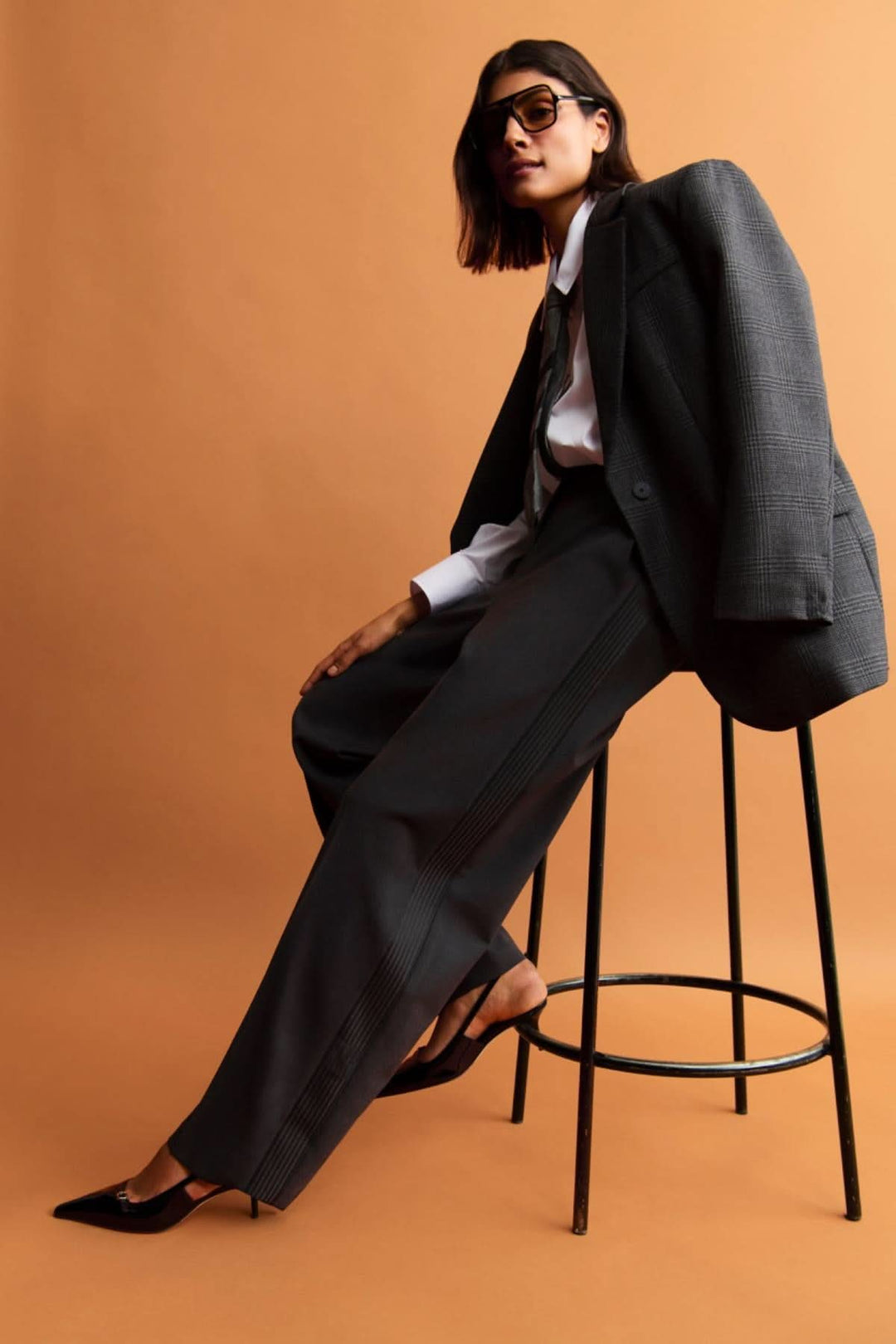 Modèle portant un pantalon large et un blazer élégant, seated sur une chaise, sur fond orange.