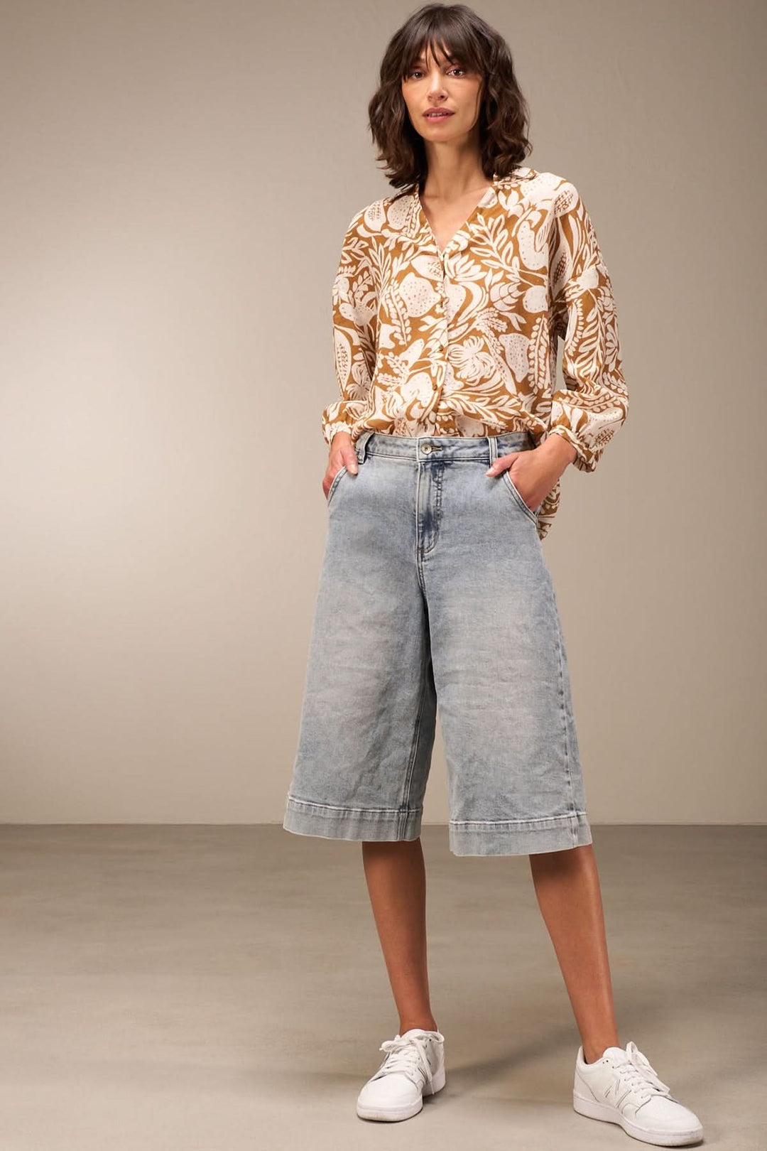 Femme portant un bermuda ample en denim délavé, chemisier floral, et baskets blanches, style détente.