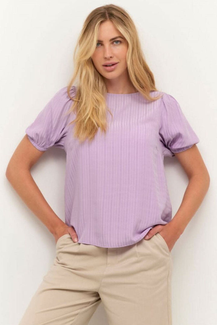 Blouse Silvia en lilas, à manches courtes, portée avec un pantalon beige, parfaite pour le printemps.