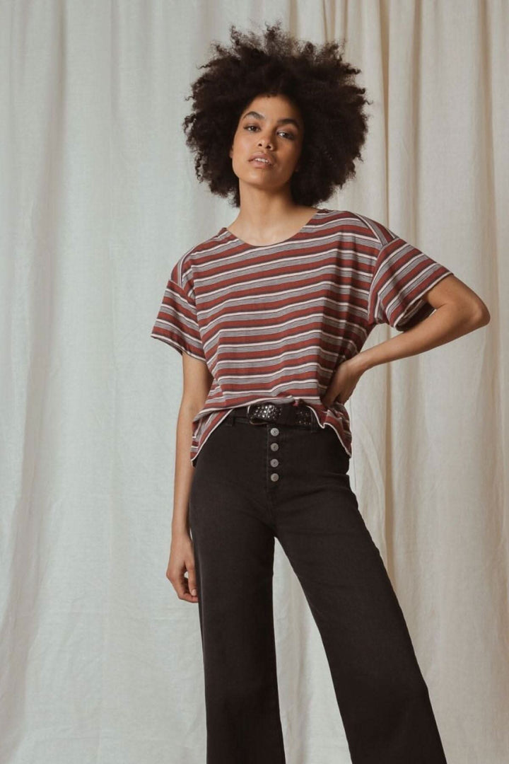 Femme stylée avec cheveux crépus, portant un t-shirt rayé et un pantalon noir devant un fond beige.