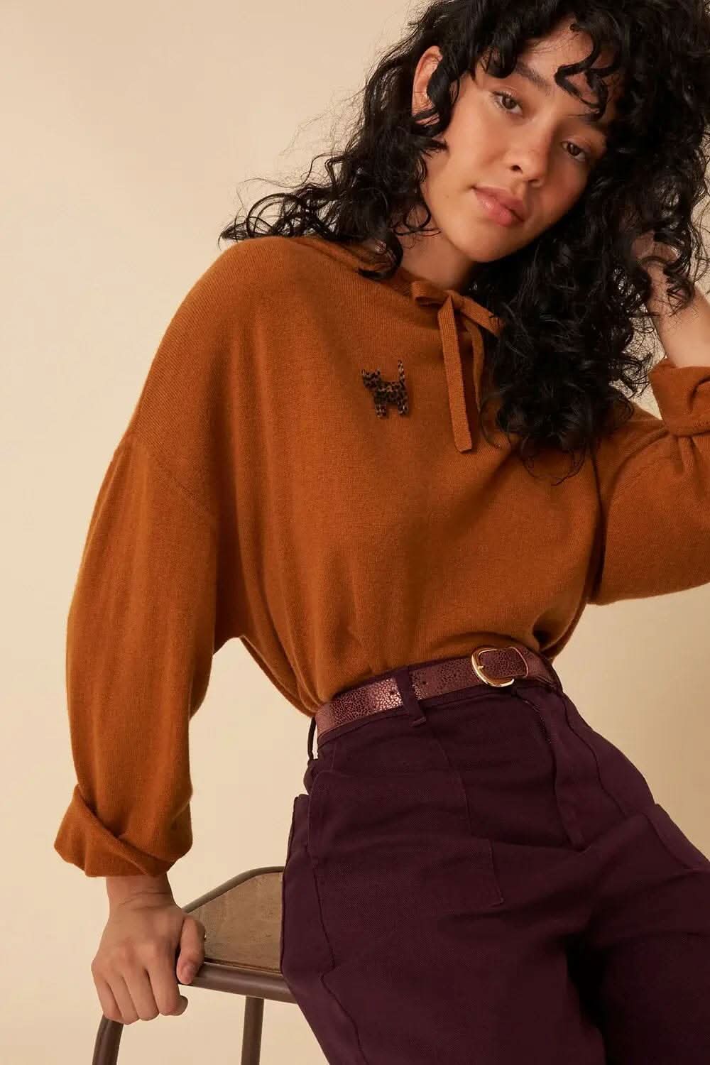 Femme portant un pull marron avec un nœud, ceinture violette et pantalon prune, style moderne et décontracté.