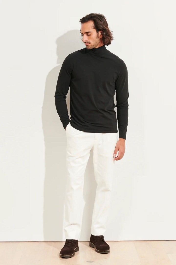 Homme portant un col roulé stretch en coton pima, pantalon blanc et chaussures brunes, devant un fond neutre.