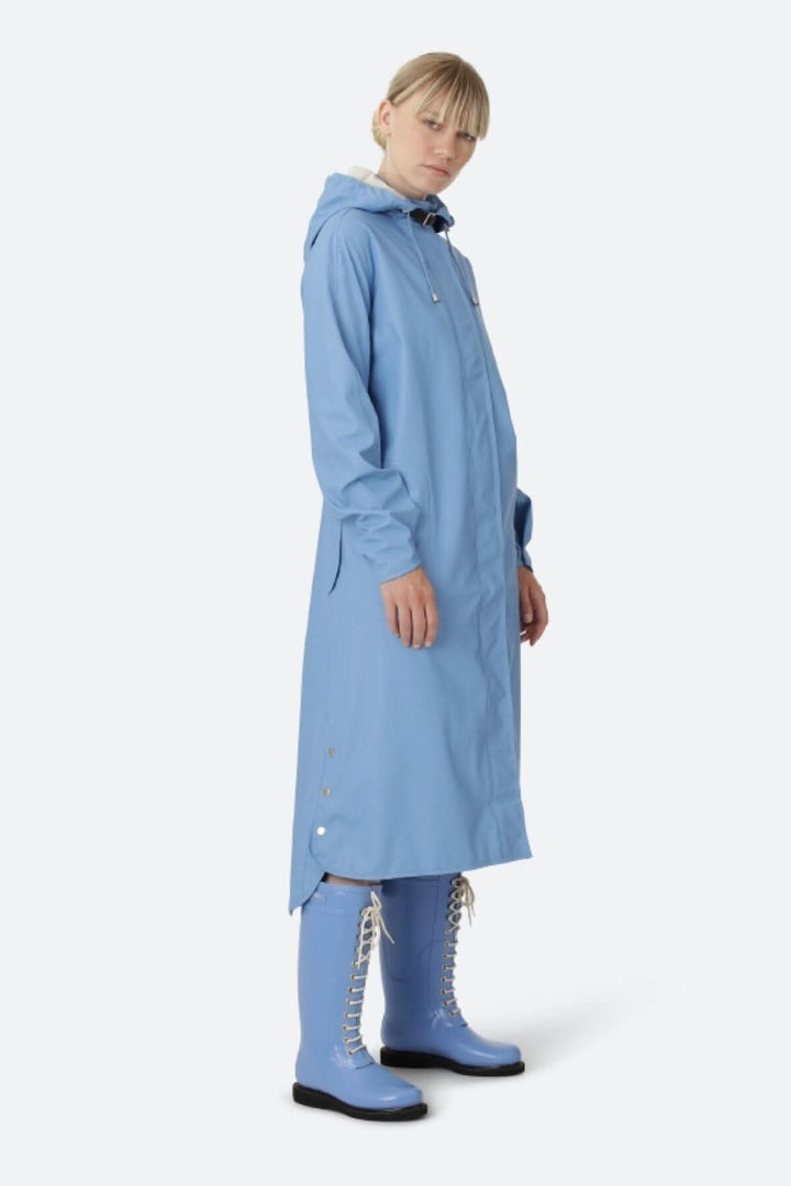 Imperméable bleu pour femme, avec capuche détachable et poches latérales, porté avec des bottes assorties.