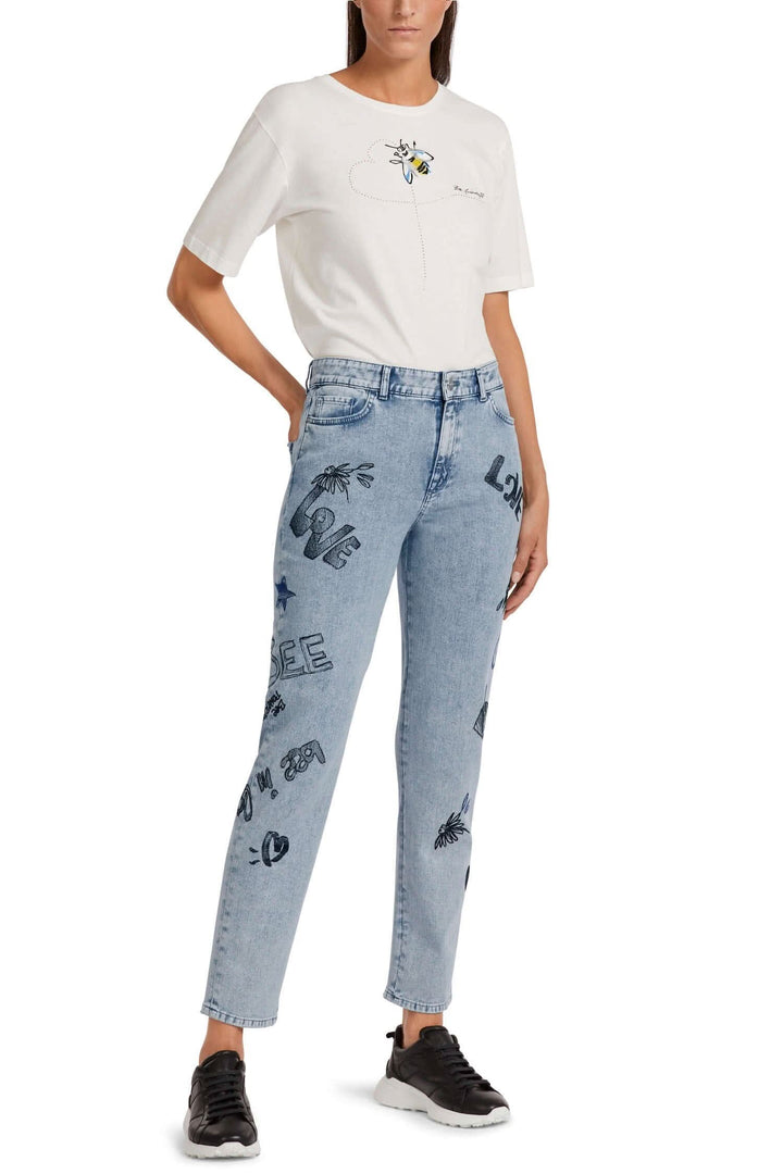 Jean tendance brodé avec écriture noire, à cinq poches, porté avec un t-shirt blanc, style décontracté.