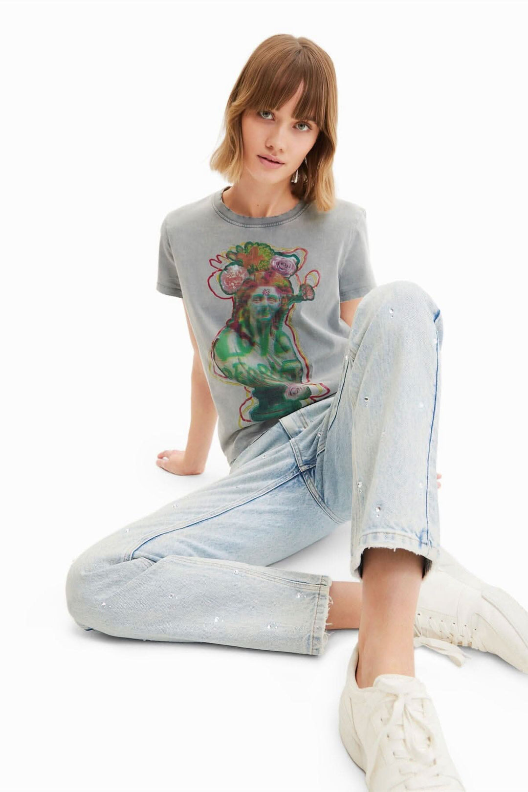Femme assise portant un t-shirt gris avec un motif coloré et un jean droit délavé avec détails en strass.