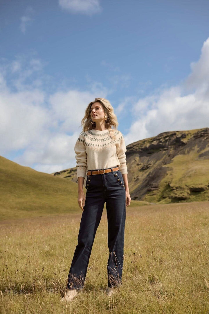Femme portant un jean ajusté et un pull beige dans un champ à la campagne, avec un paysage montagneux en arrière-plan.