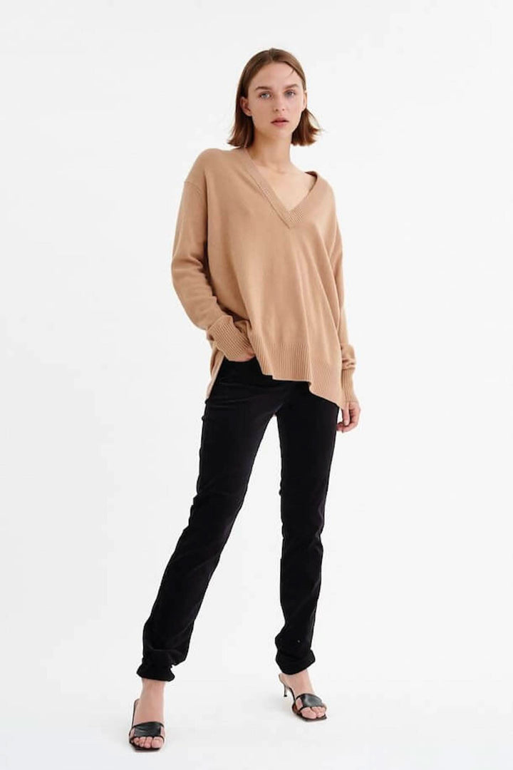 Femme portant un pull beige, debout avec un pantalon noir, look décontracté et moderne.