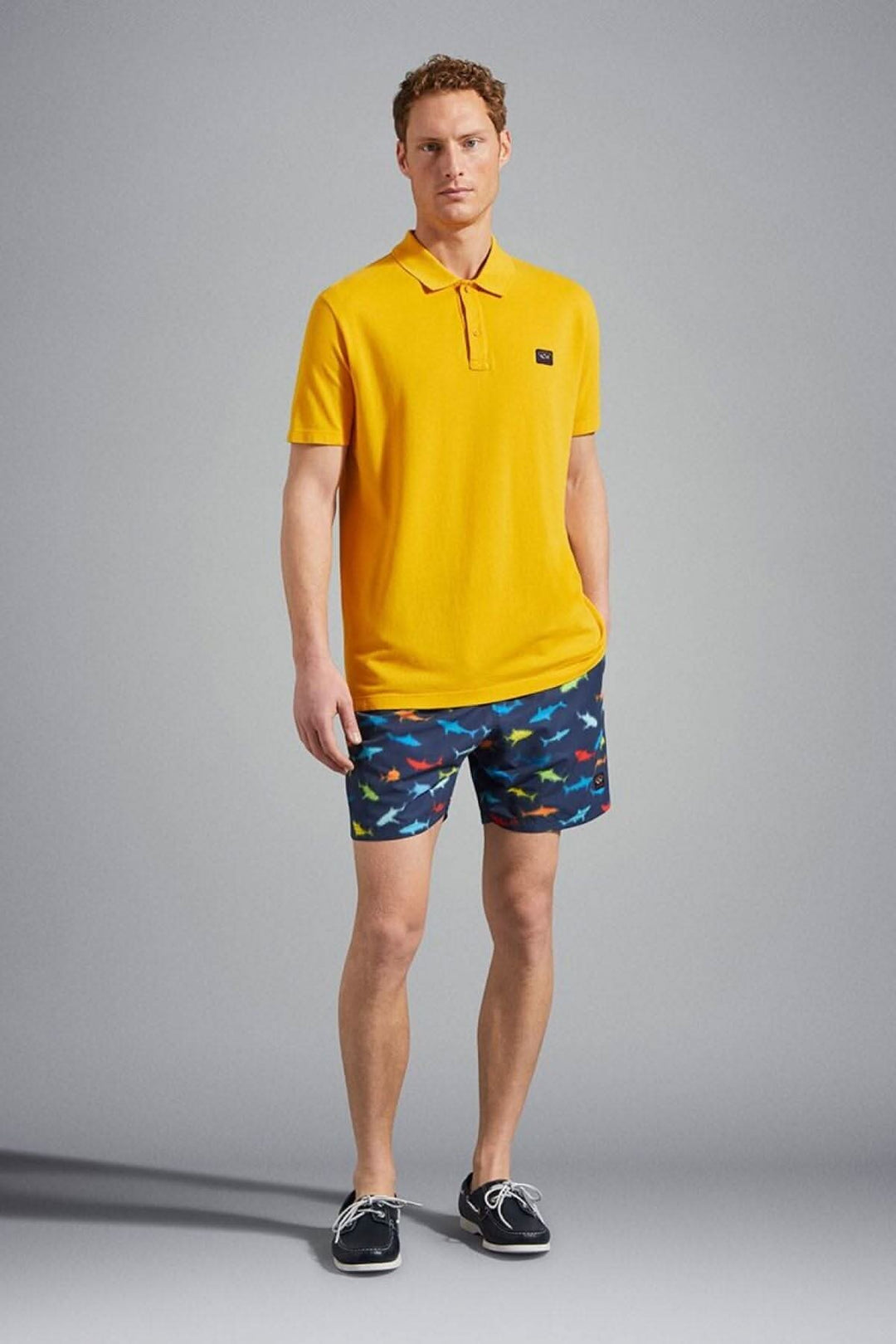 Homme portant un maillot de bain avec imprimé requin et un polo jaune de la marque Paul and Shark.