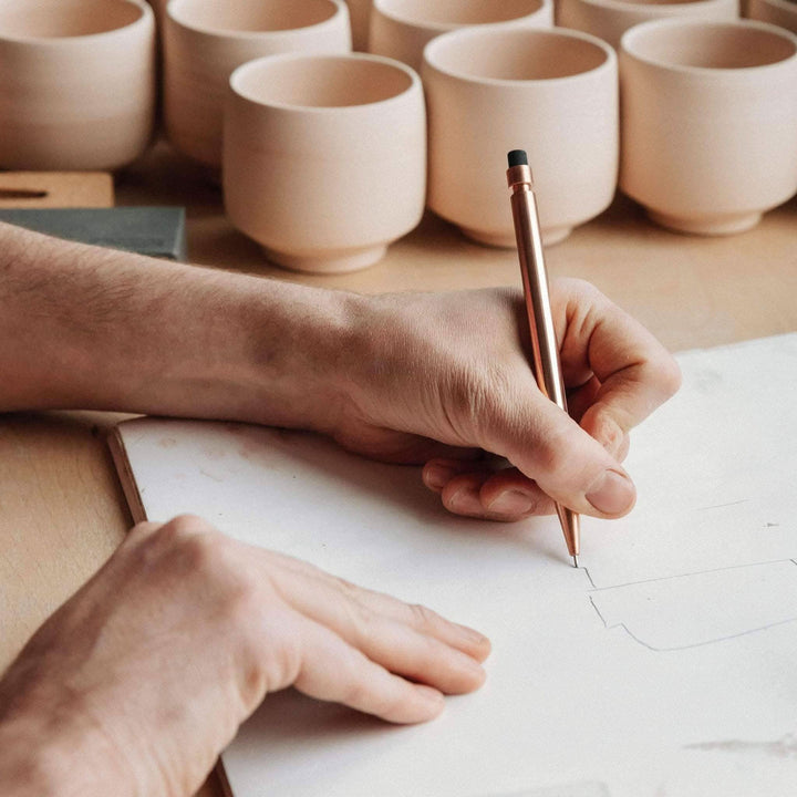 Un artisan utilise un crayon Modern Fuel pour dessiner sur une planche à côté de tasses en céramique.
