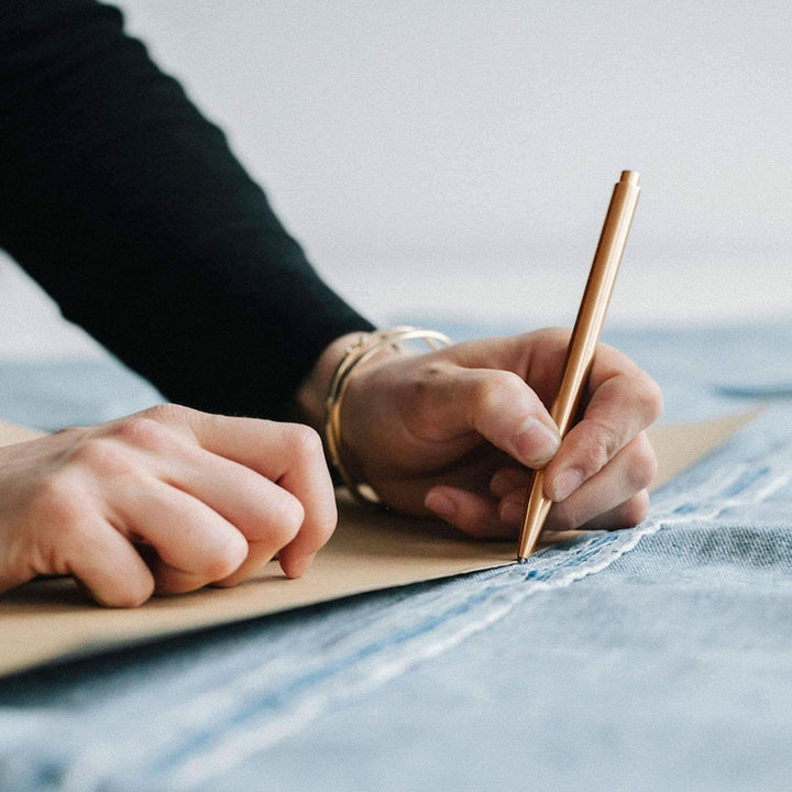 Personne utilisant un crayon Modern Fuel doré pour dessiner sur du papier situé sur un denim.