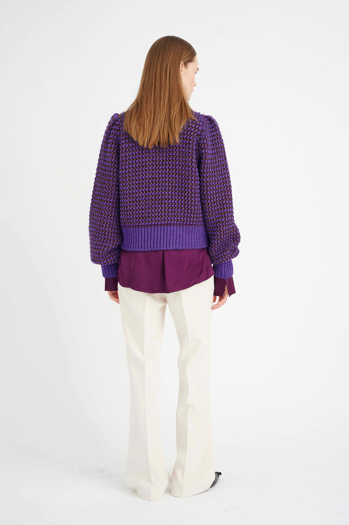 Femme de dos avec un pull violet à manches bouffantes et un pantalon clair, sur fond blanc.