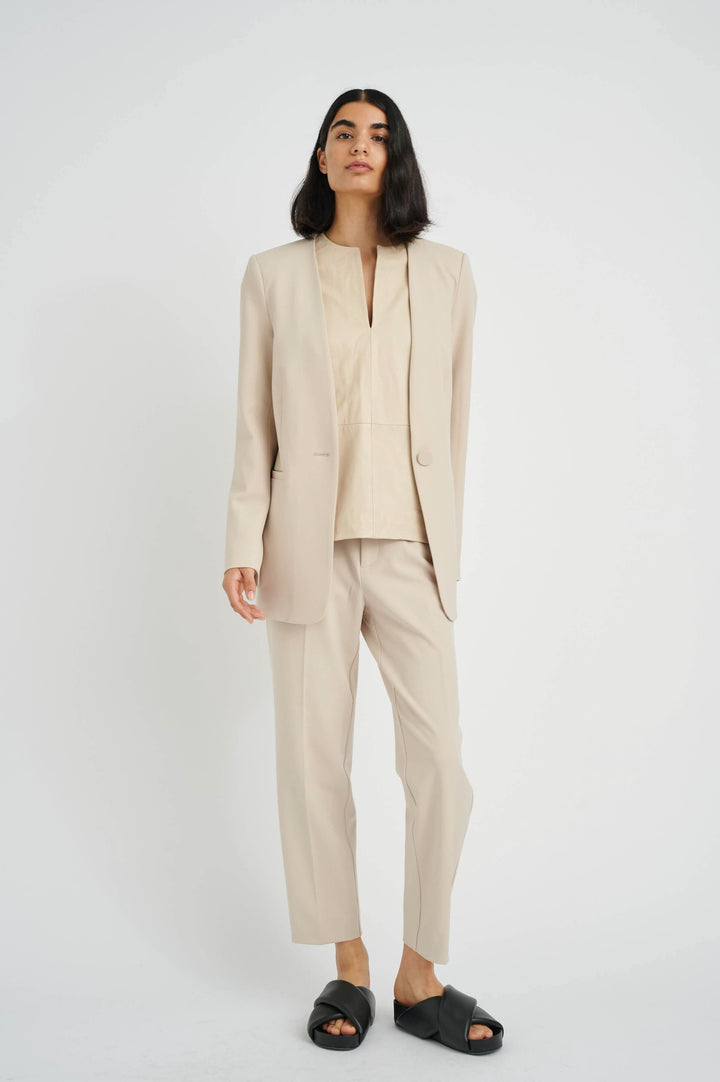 Femme portant un ensemble de pantalon et veste beige élégant avec des sandales noires, sur fond blanc.