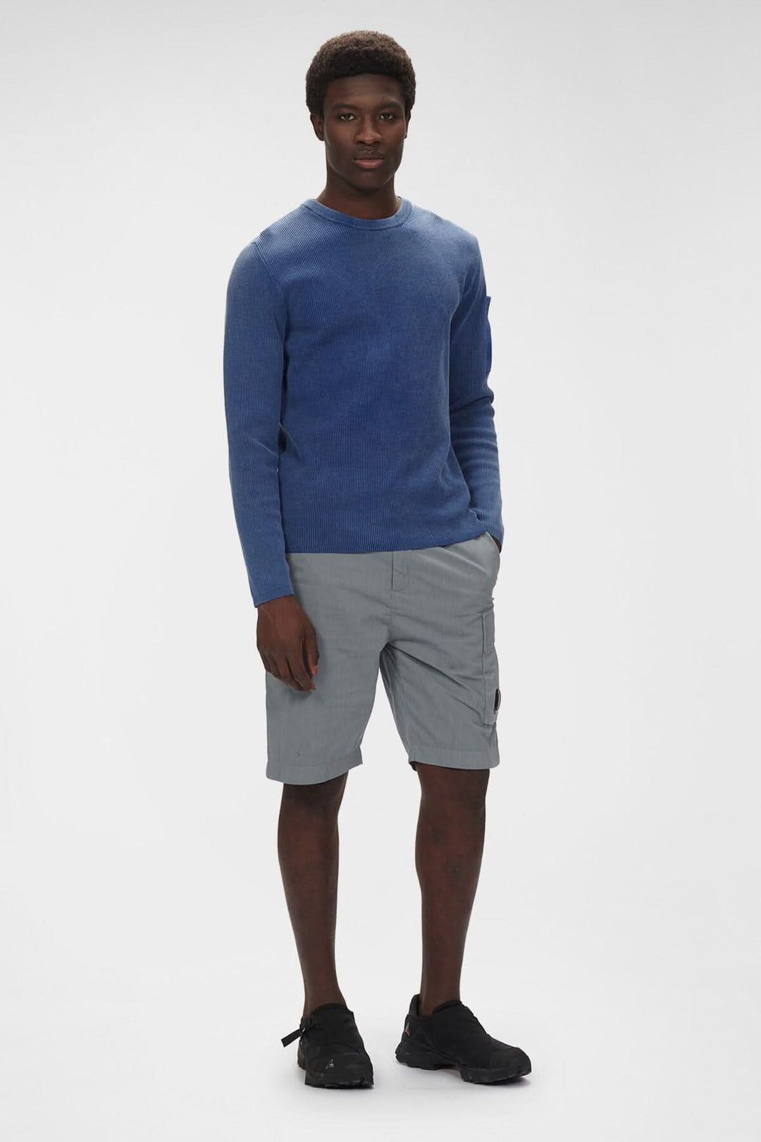 Homme portant un pull bleu et un short cargo gris, avec des poches, sur un fond neutre.